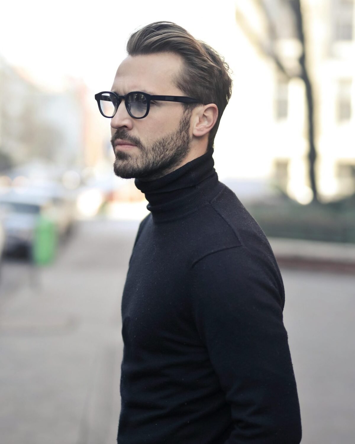 A fashionable man in a black top and glasses poses in an urban street with parked cars.